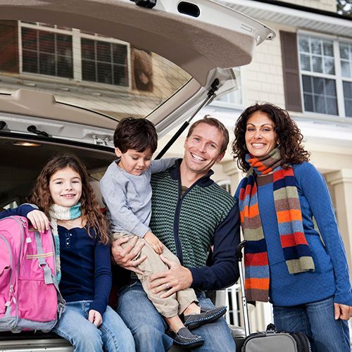 Family of four smiling, near an open car trunk; girl with backpack, boy held, house in background.